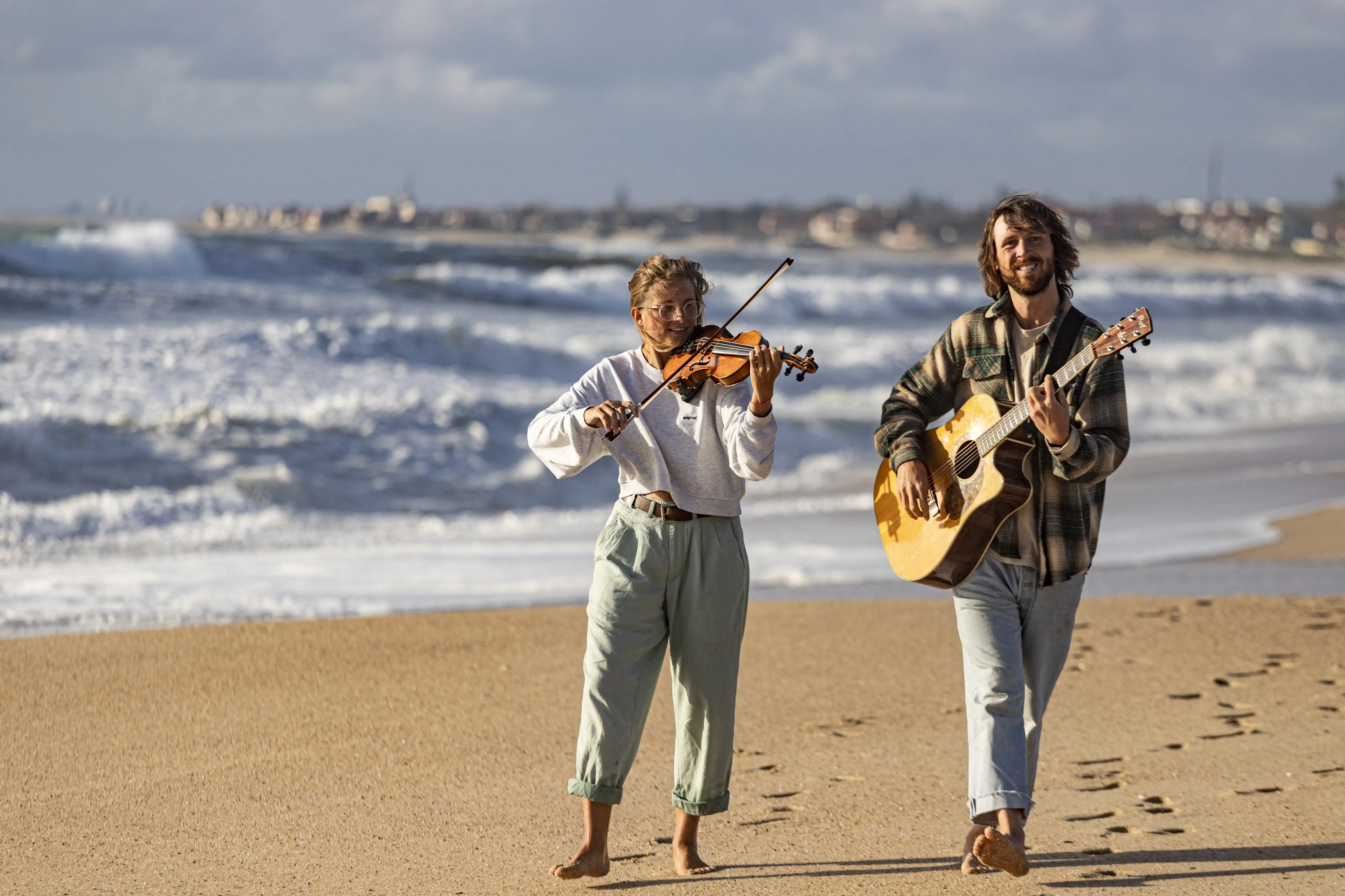 Anka und René laufen am Strand und spielen auf der Geige und auf der Gitarre.
      Die Sonne scheint und im Hintergrund brechen weiße Wellen.